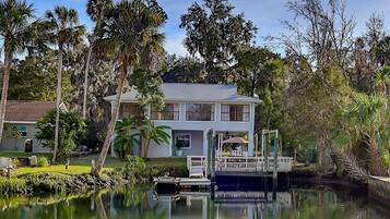 A two-story house with a dock extending into a calm body of water, surrounded by lush greenery and tall trees.