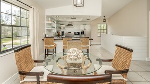 Un comedor con una mesa de vidrio rodeada de sillas de mimbre, una cocina al fondo y vistas al exterior.