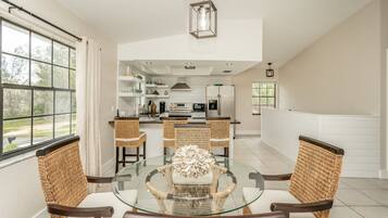 A dining area with a glass table surrounded by wicker chairs, a kitchen in the background, and a view of the outdoors.