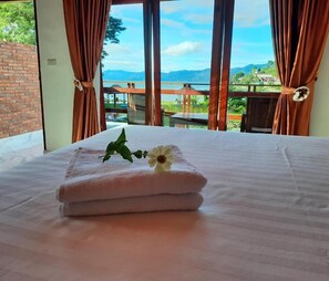 A hotel room with a bed, a window with curtains, and a view of a mountainous landscape. - Juma Cottages (Samosir)