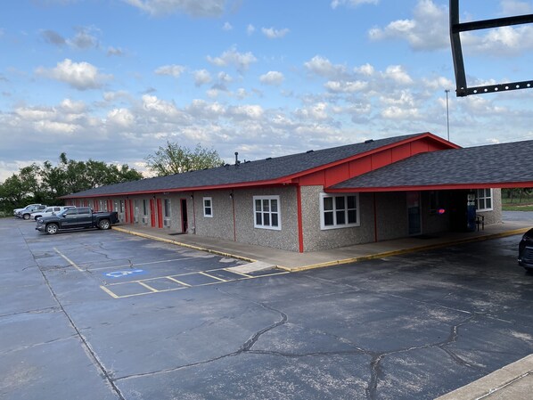 Un motel à l’extérieur rouge et gris, doté d’un parking couvert et surplombant un ciel dégagé parsemé de nuages.