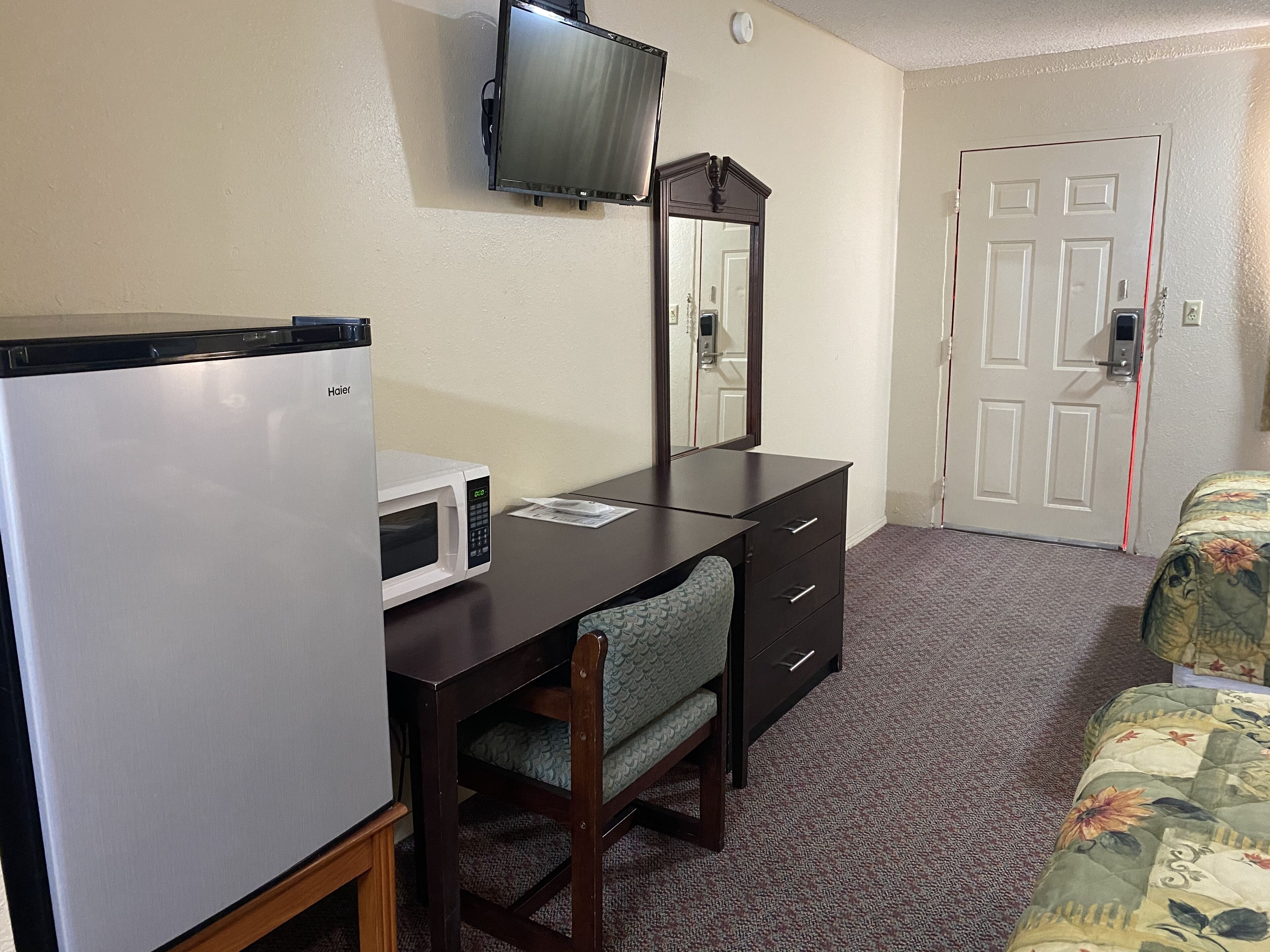 Habitación de hotel compacta con kitchenette, televisor de pantalla plana montado en la pared, escritorio de madera con silla, espejo grande y puerta con línea de seguridad roja.