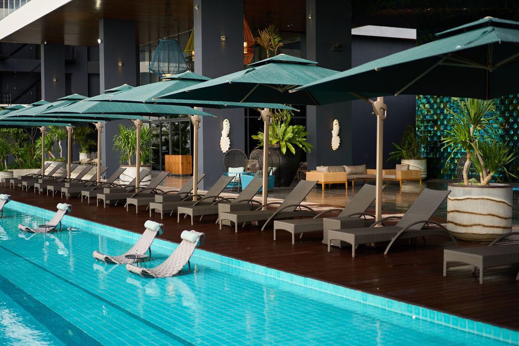 Un área de piscina con sillas de descanso y sombrillas, rodeada de plantas en macetas y la fachada de un edificio moderno.