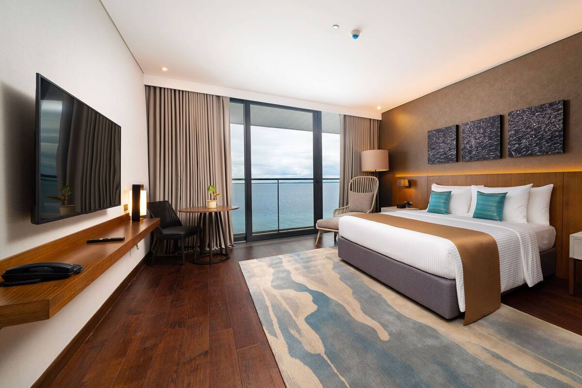Ett modernt hotellrum med en stor säng, ett skrivbord, en stol, en TV och utsikt över havet.