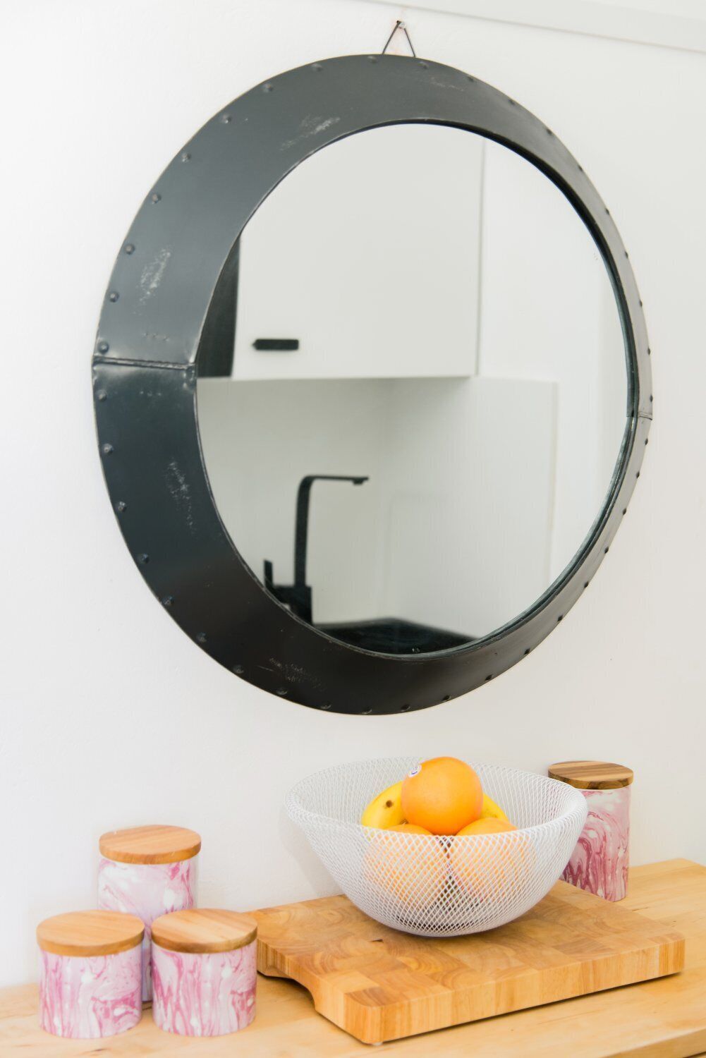 A round mirror on a wall, a wooden cutting board, a white mesh basket with oranges, and three cylindrical containers with wooden lids.