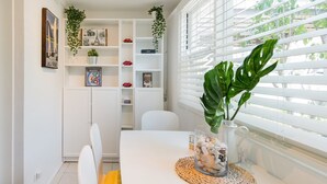 Un coin repas avec une table blanche, des chaises et une fenêtre avec des stores. On y trouve des étagères ornées d’objets décoratifs et de plantes.