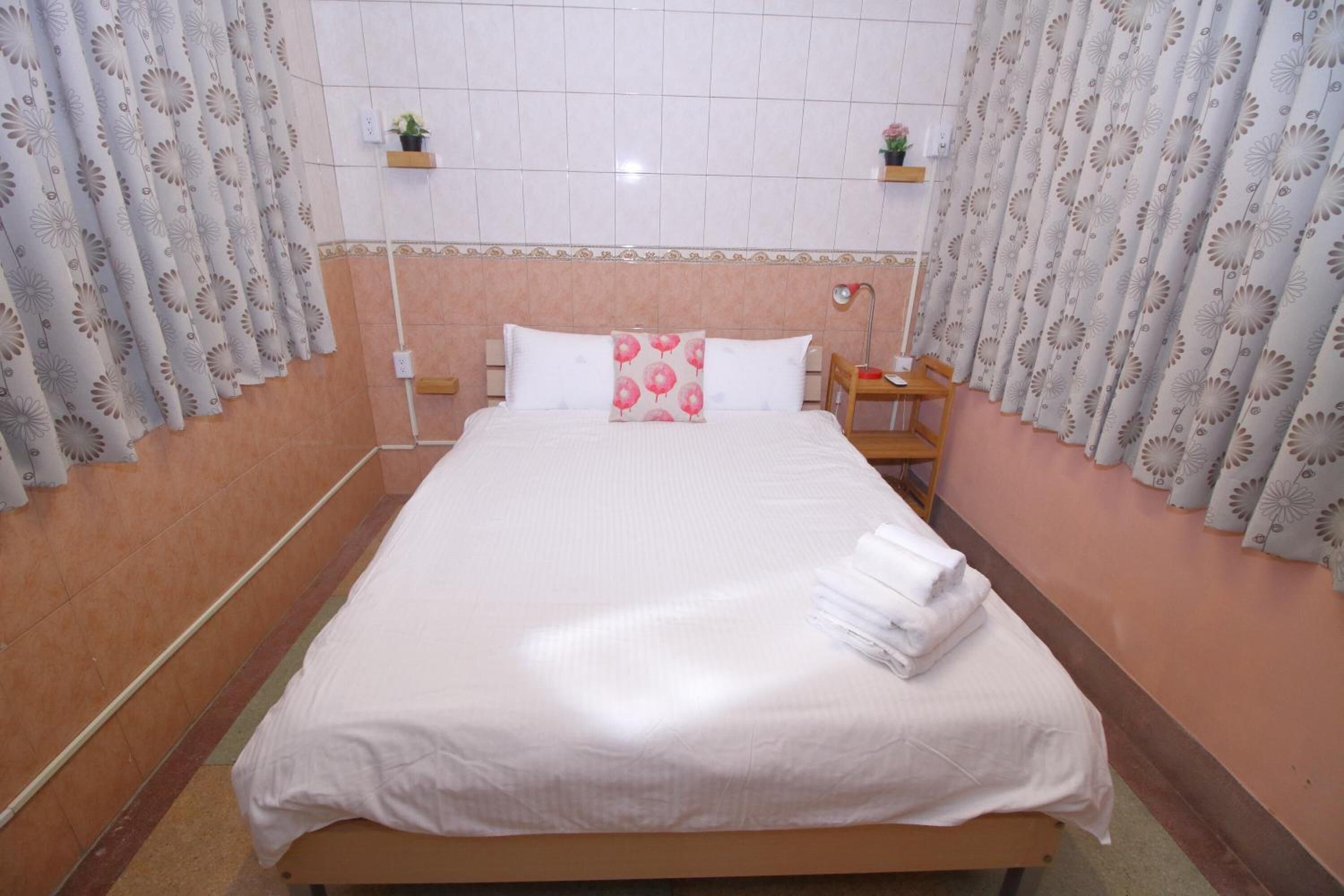 Un lit bien fait, avec du linge de lit blanc et un coussin rose, une table de chevet en bois avec une lampe, et une petite étagère avec une plante.