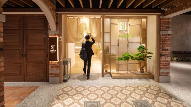 Entrada de un hotel con fachada de madera, mostrador de recepción y una planta en maceta.