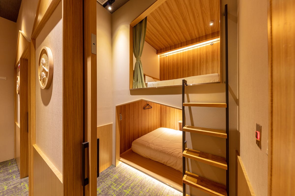 Une petite pièce avec un lit superposé, une échelle en bois, un petit miroir et un luminaire au-dessus du lit.