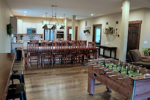 A spacious living room with a wooden dining table surrounded by chairs, a foosball table, and a kitchen area with modern appliances.