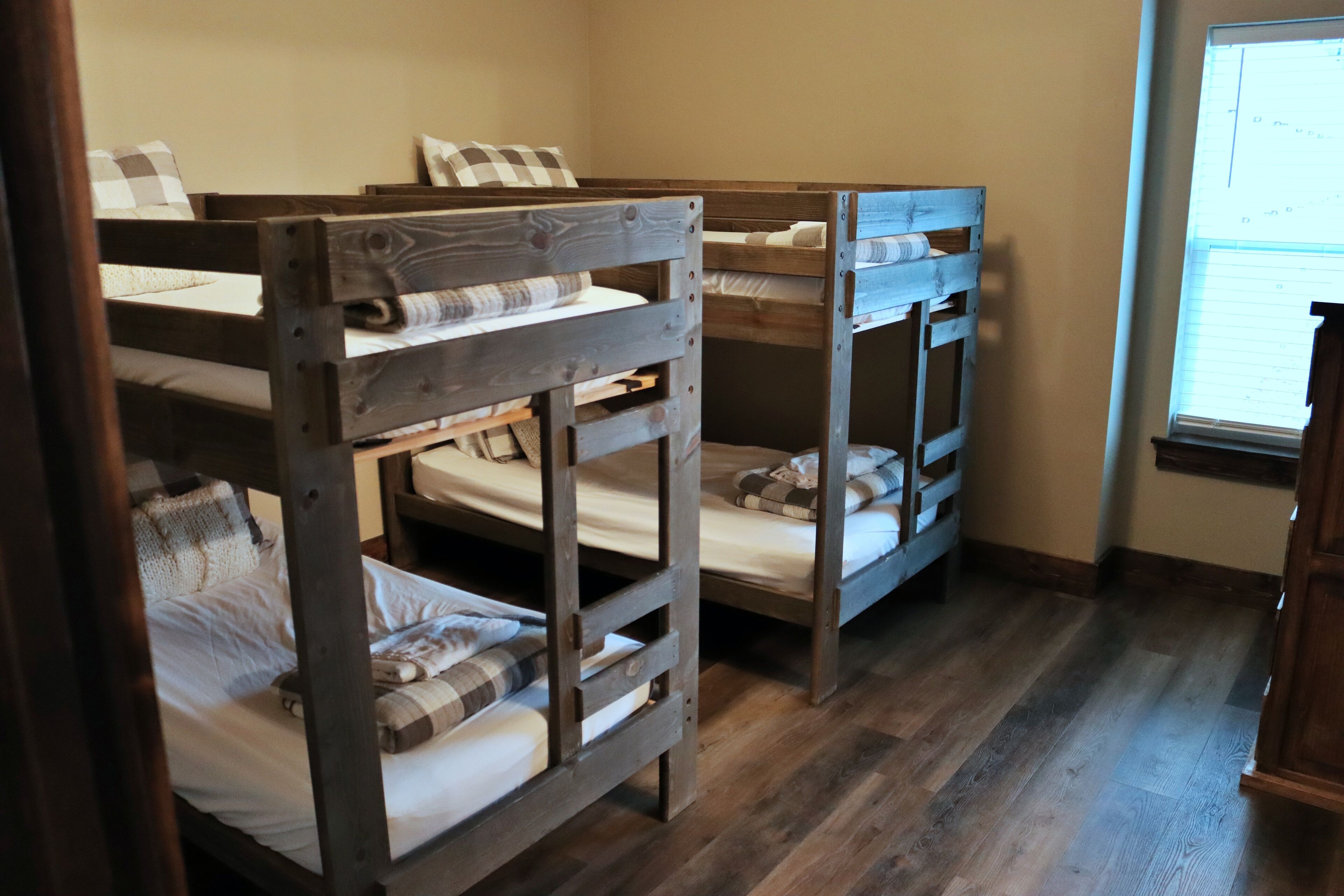 A room with three bunk beds, wooden flooring, and a window with blinds.