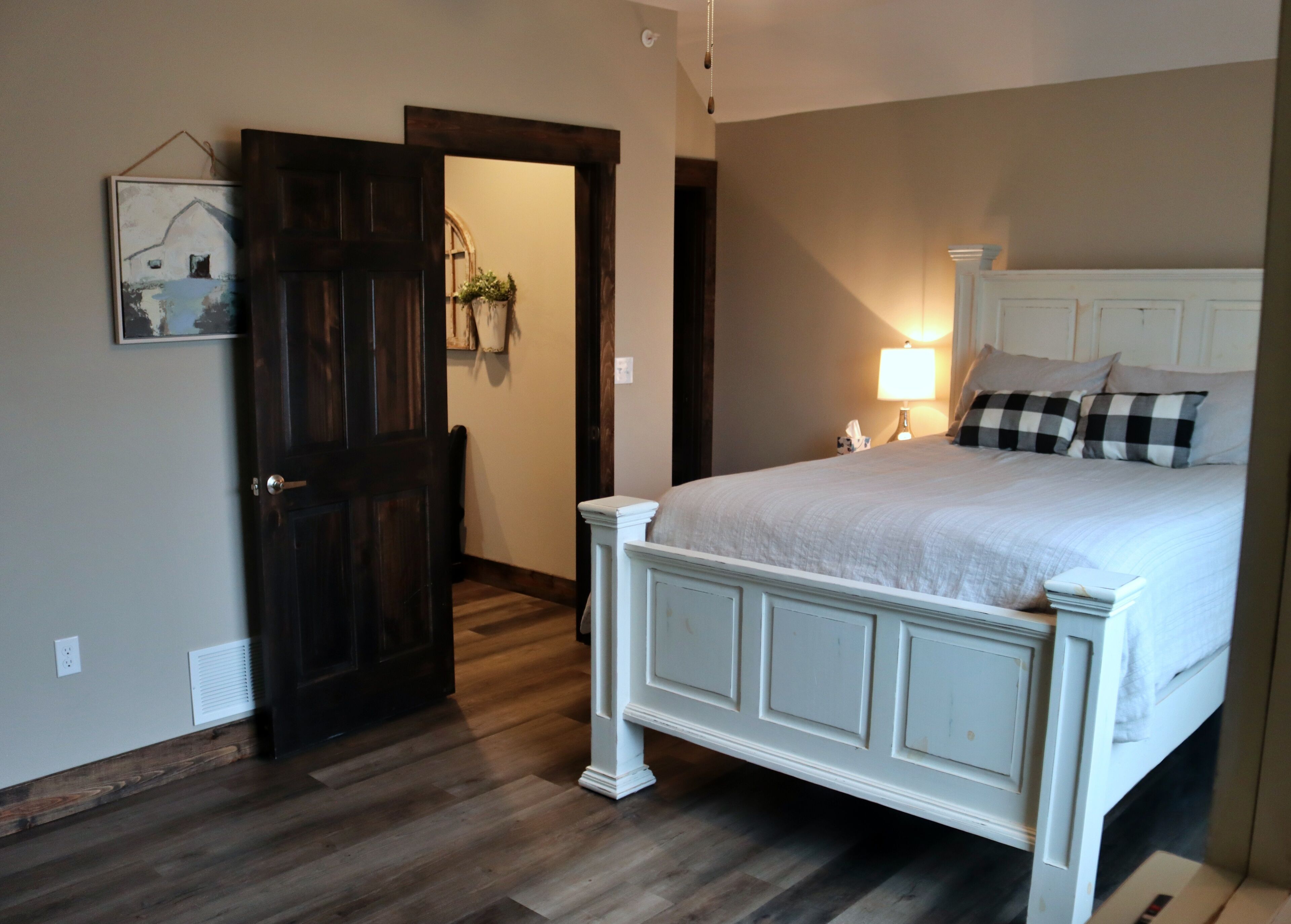 A bedroom with a large bed, a wooden door, a wall-mounted picture, and a bedside lamp.