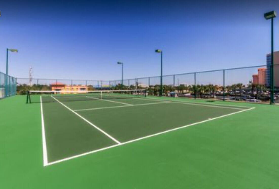 Un court de tennis en plein air, sous un ciel dégagé, entouré d’une clôture et éclairé par des lampadaires.