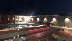 A pool area with lounge chairs and umbrellas, a wooden deck, and a building with illuminated windows. - Mid Century Modern Home || 28 day minimum  (Palm Springs)