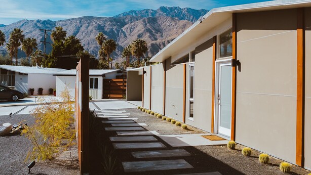 A modern building with a paved walkway leading to a garage, surrounded by palm trees and mountains in the background. - Mid Century Modern Home || 28 day minimum  (Palm Springs)