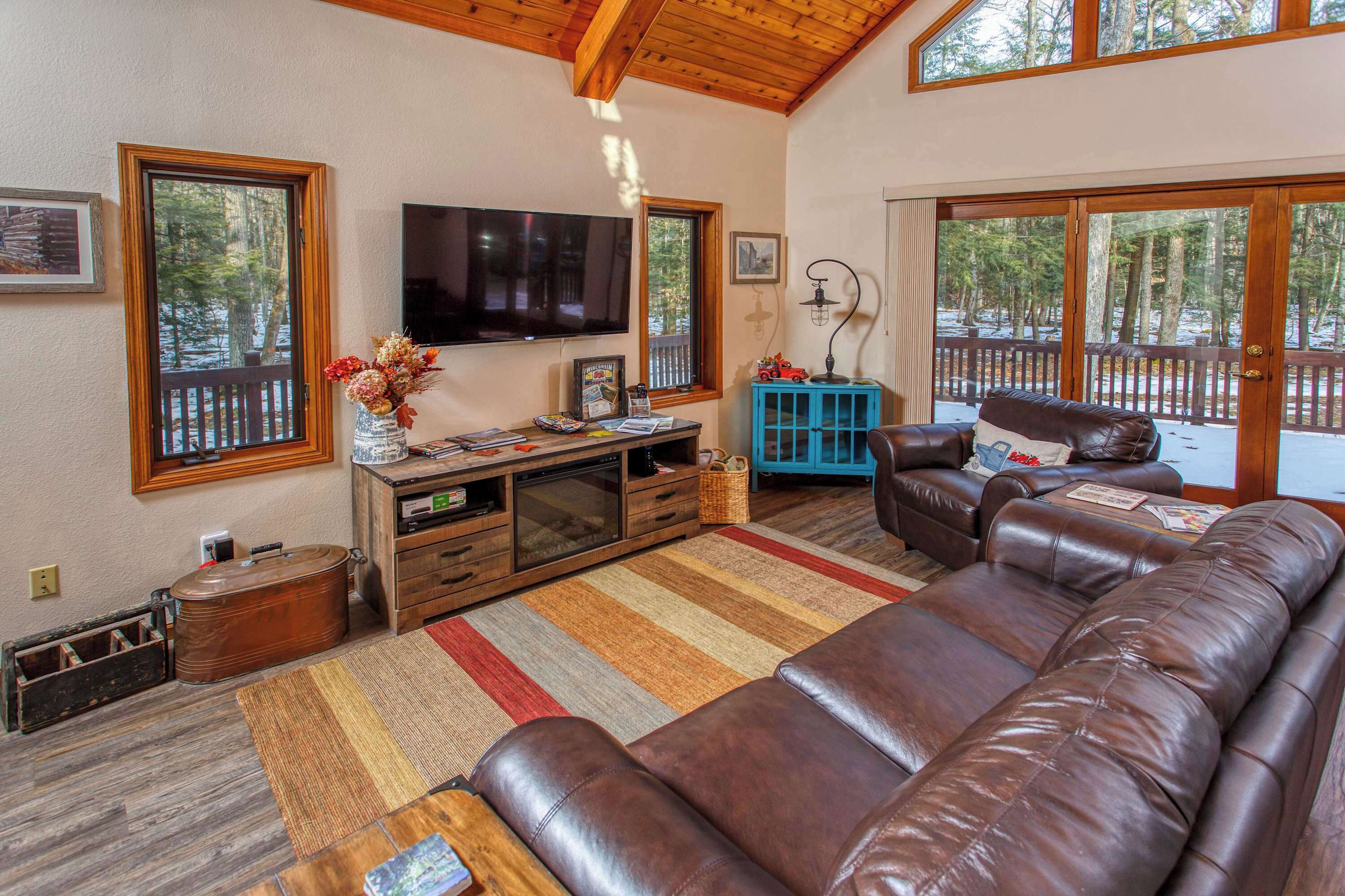 Una sala de estar con un amplio sofá de cuero marrón, un televisor de pantalla plana, un mueble de entretenimiento de madera y vistas a una terraza a través de grandes ventanales.