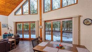 A spacious living area with a wooden ceiling, large windows, a dining table with placemats, a sofa, and a view of a snowy deck. - Dog-Friendly, Secluded Cabin in the Woods (Egg Harbor)