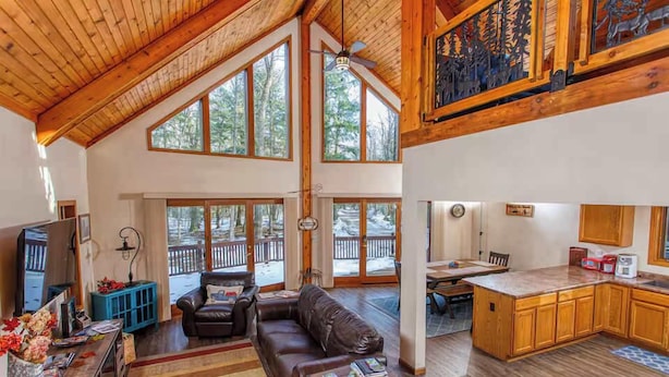 A spacious living area with a large window, a fireplace, and a kitchen with wooden cabinets. - Dog-Friendly, Secluded Cabin in the Woods (Egg Harbor)