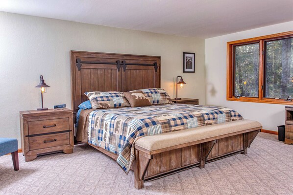Quarto com cama de madeira, duas mesas de cabeceira, um banco e janela com vista para árvores.