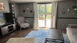 Una sala de estar con televisor, un sillón cómodo, un sofá, una mesa de centro y un ventanal corredero que da a un patio exterior.