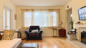 Un salon comprenant un canapé, un fauteuil, un plancher en bois, une cheminée, un téléviseur et une fenêtre avec des rideaux légers.