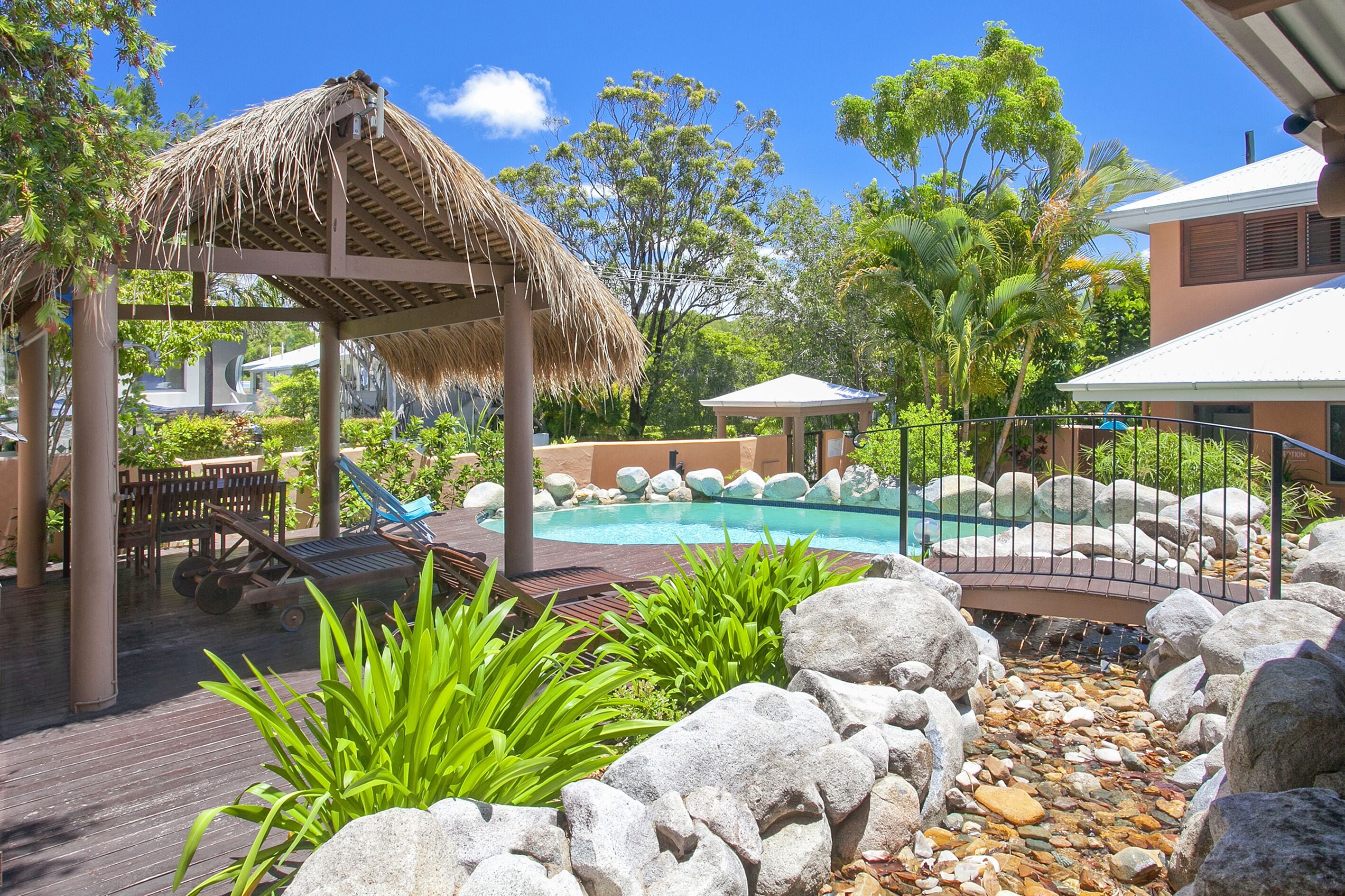 A pool area with lounge chairs, a thatched gazebo, and a rocky landscape.