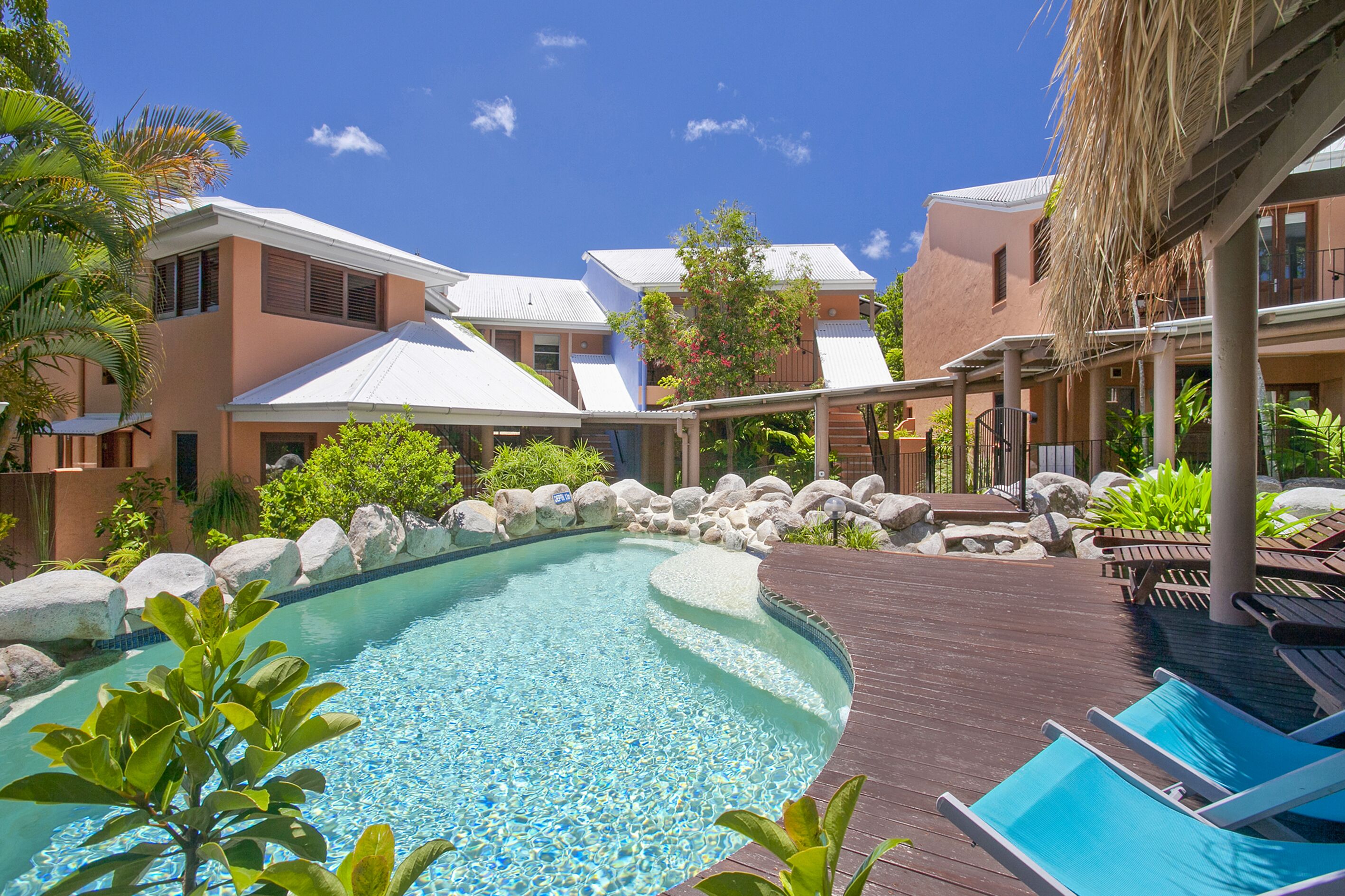 A pool area with a wooden deck, lounge chairs, and a clear blue pool surrounded by rocks and greenery.