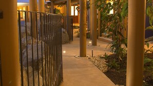 Une allée couverte, bordée de colonnes, avec une clôture en métal et des plantes.