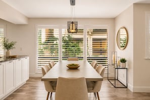 A dining area with a long table, chairs, a bowl of food, a pendant light, and a view through large windows. - PGA National Home on Golf Course by Guaranteed Rental™ (Palm Beach Gardens)
