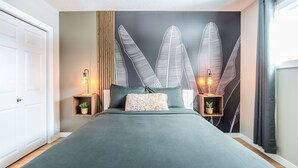 A modern bedroom with a large bed, two bedside tables with lamps, a wall-mounted shelf with plants, and a leaf-patterned wall. - 5BR Bungalow: AC|Arcade|Foosball|Walk to WEM (Edmonton)