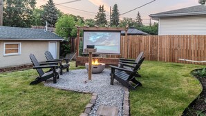 A backyard with a fire pit, two Adirondack chairs, and a projector screen displaying a mountain scene. - 5BR Bungalow: AC|Arcade|Foosball|Walk to WEM (Edmonton)
