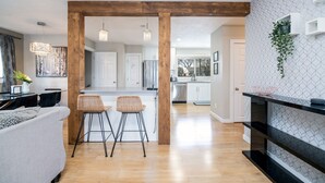 A modern kitchen with wooden beams, a dining area with a sofa and two bar stools, and a black shelving unit against a patterned wall. - 5BR Bungalow: AC|Arcade|Foosball|Walk to WEM (Edmonton)