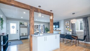 A modern kitchen with white cabinets, a central island, and a dining area with a glass table and black chairs. - 5BR Bungalow: AC|Arcade|Foosball|Walk to WEM (Edmonton)