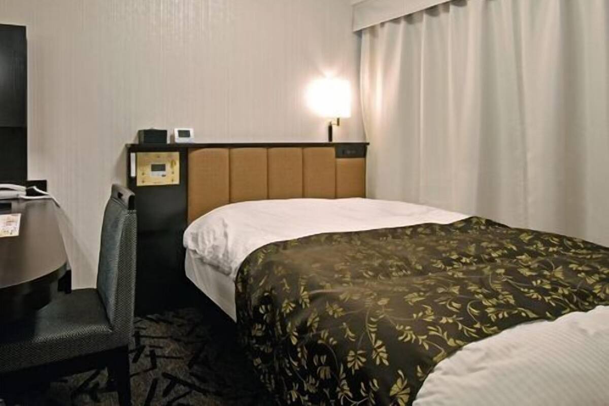 A hotel room with a bed, a desk, a chair, a nightstand, and a lamp.