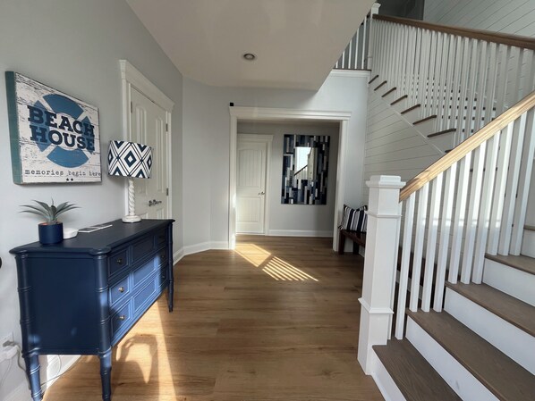 A hallway with a staircase, wooden floors, a blue dresser, and a 'Beach House' sign.