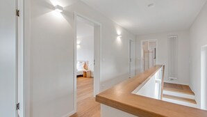 A modern interior with a wooden staircase, white walls, and a visible bedroom with a bed and nightstand.