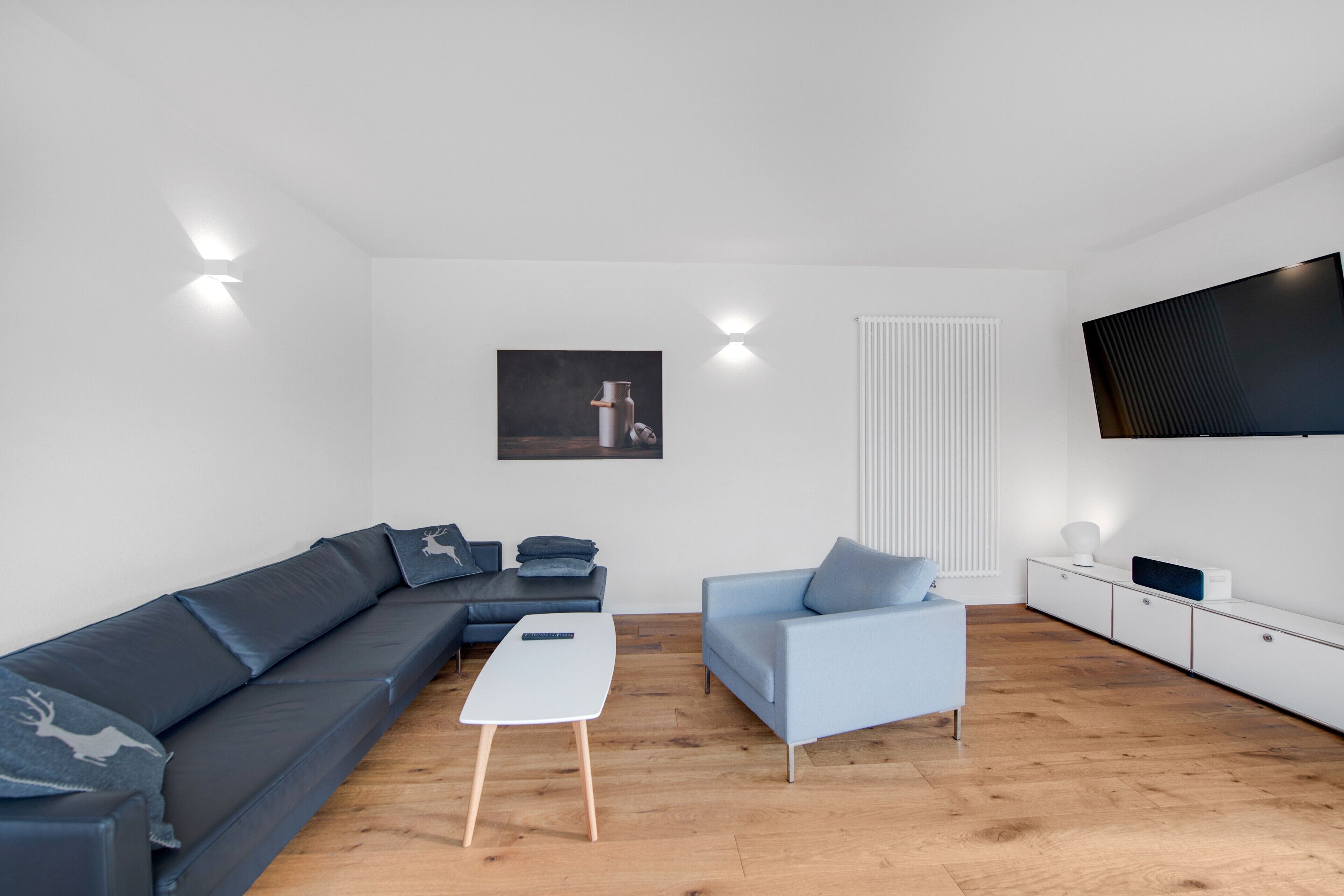 Een moderne woonkamer met een modulaire bank, een lichtblauwe fauteuil, een witte salontafel en een flatscreen televisie aan de muur.