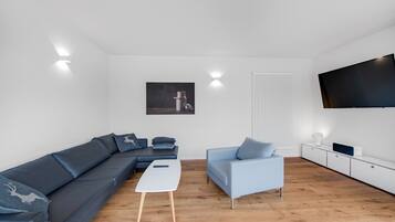 Een moderne woonkamer met een modulaire bank, een lichtblauwe fauteuil, een witte salontafel en een flatscreen televisie aan de muur.