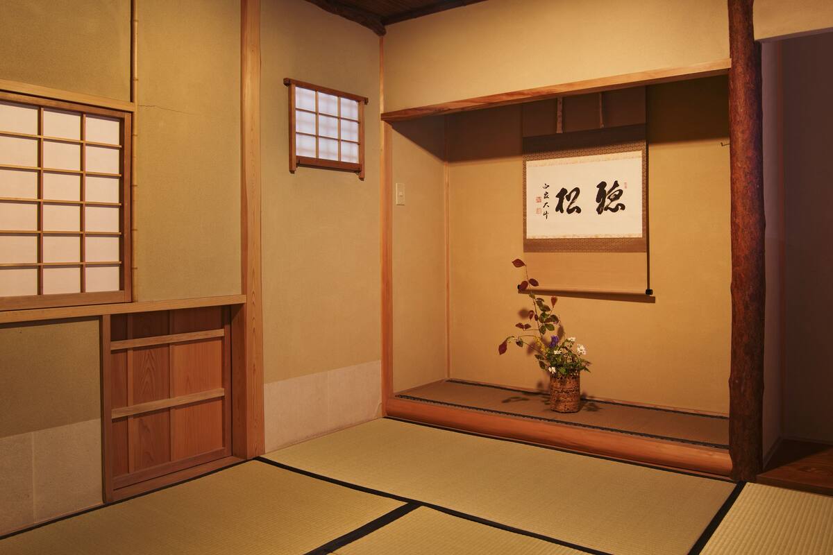 A traditional Japanese room with tatami flooring, a shoji screen, a small window, and a calligraphy piece on the wall.