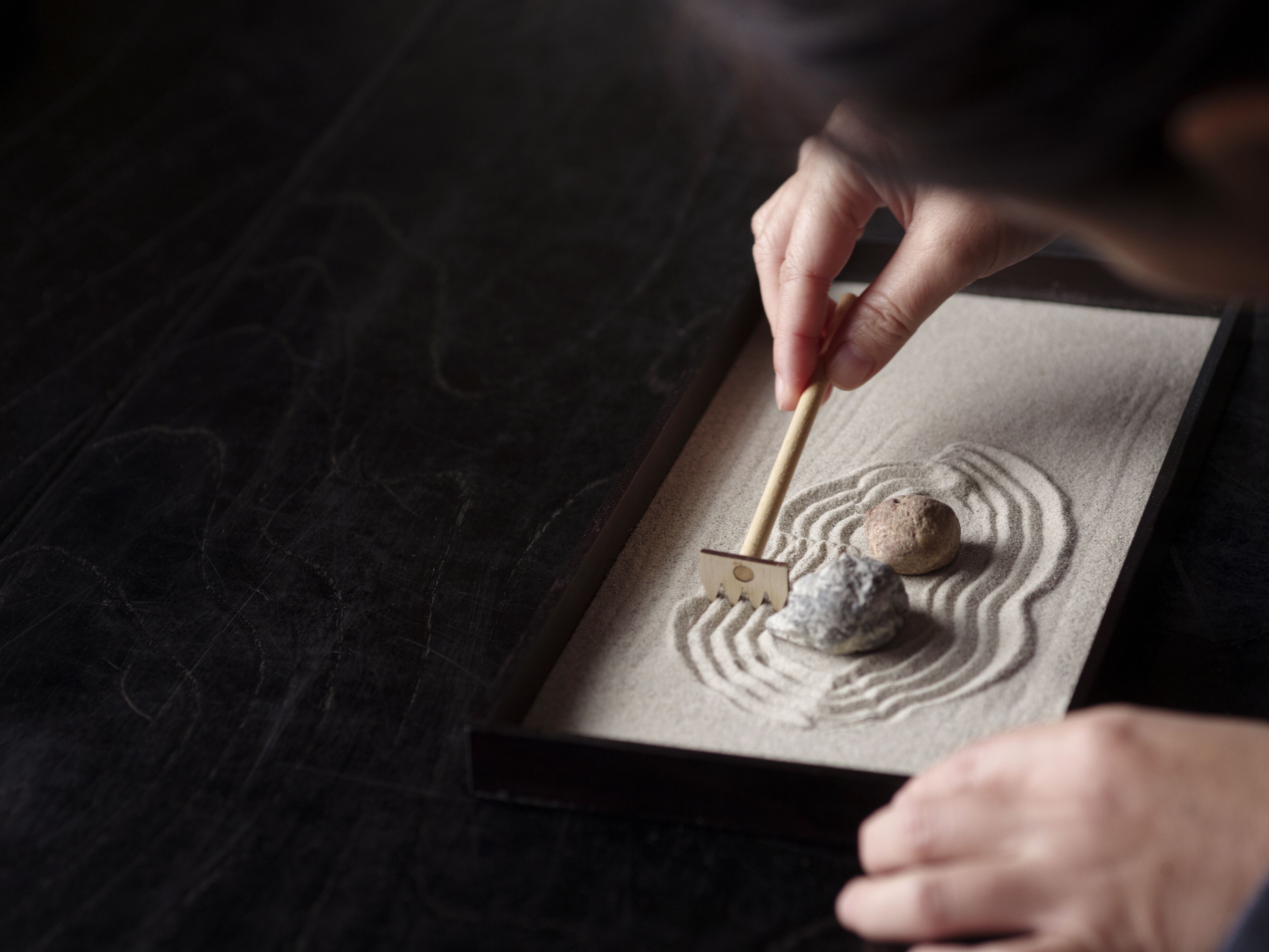 A person using a wooden tool to rake sand in a rectangular container, creating patterns.