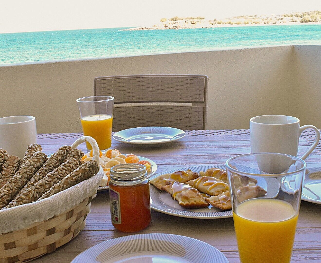 A table set with breakfast items including bread, pastries, and drinks, overlooking a beach.