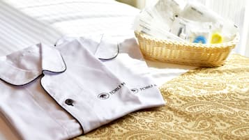 Una camisa blanca doblada con cuello y botón negros, sobre una cama con colcha estampada en tonos dorado y beige. Se ve una cesta de mimbre con artículos de aseo en el fondo.