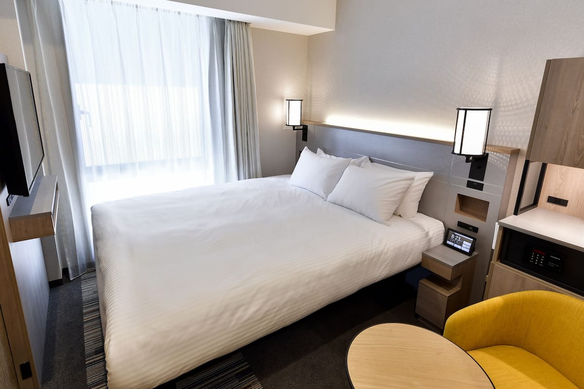 A hotel room with a large bed, bedside tables, a TV, a desk with a chair, and a window with curtains.