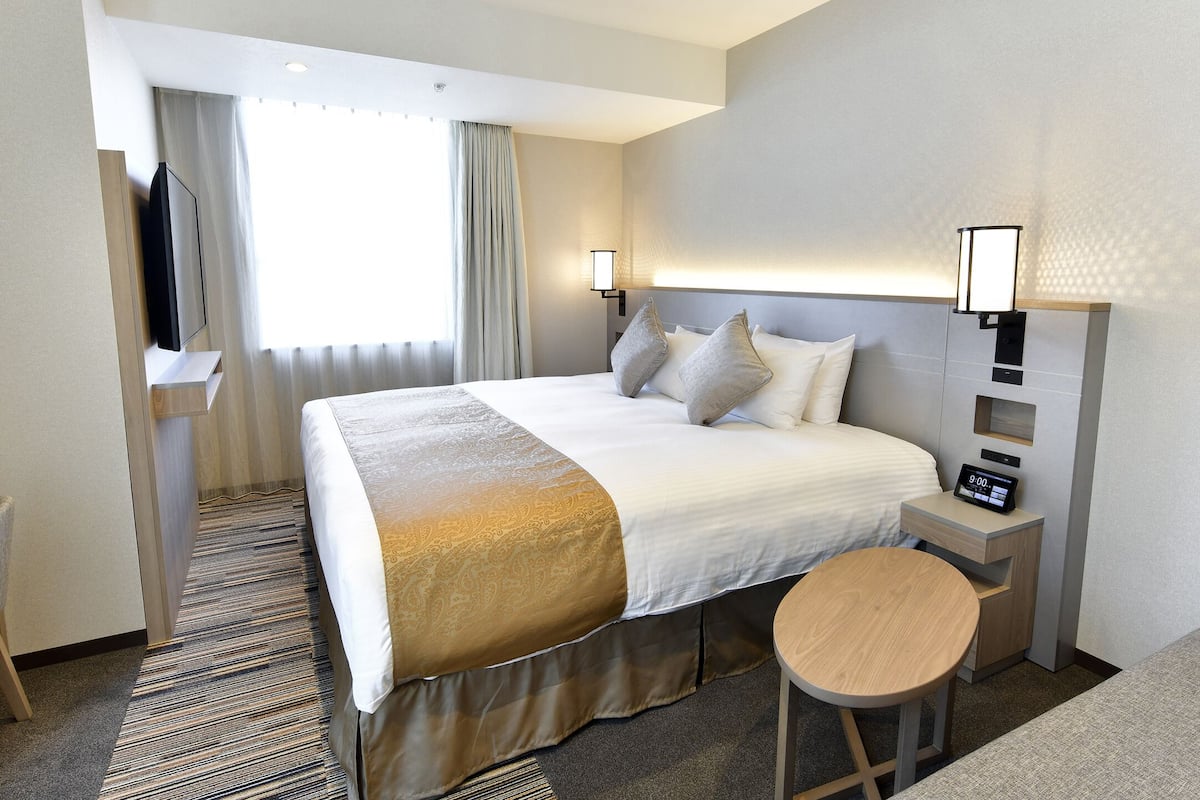 A hotel room with a bed, bedside tables, a desk, a chair, and a small stool.