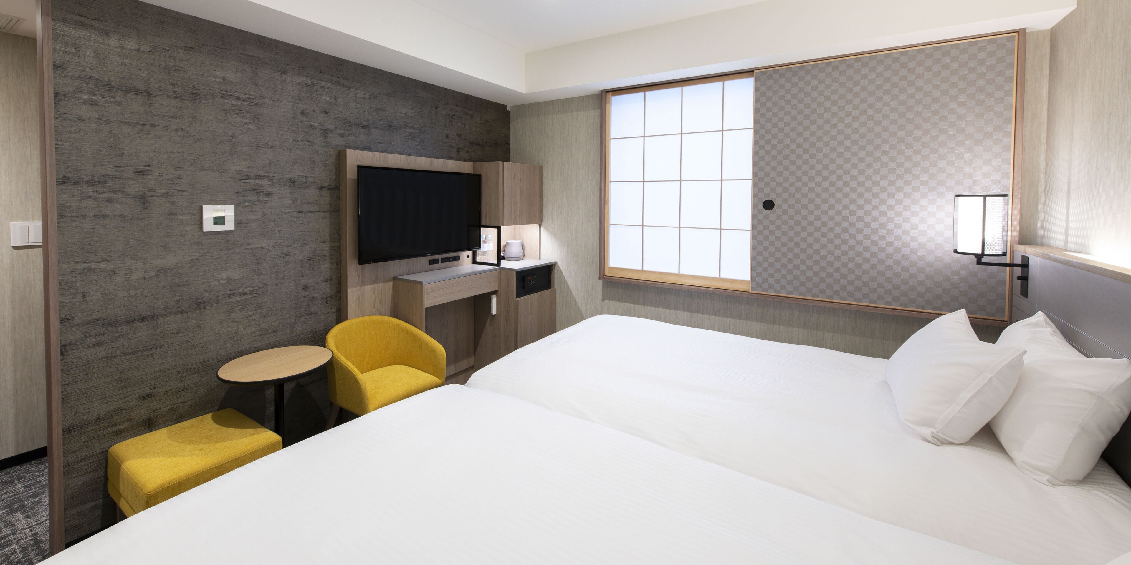 A hotel room with a large bed, a small desk with a flat-screen TV, a yellow armchair, and a window with a view.
