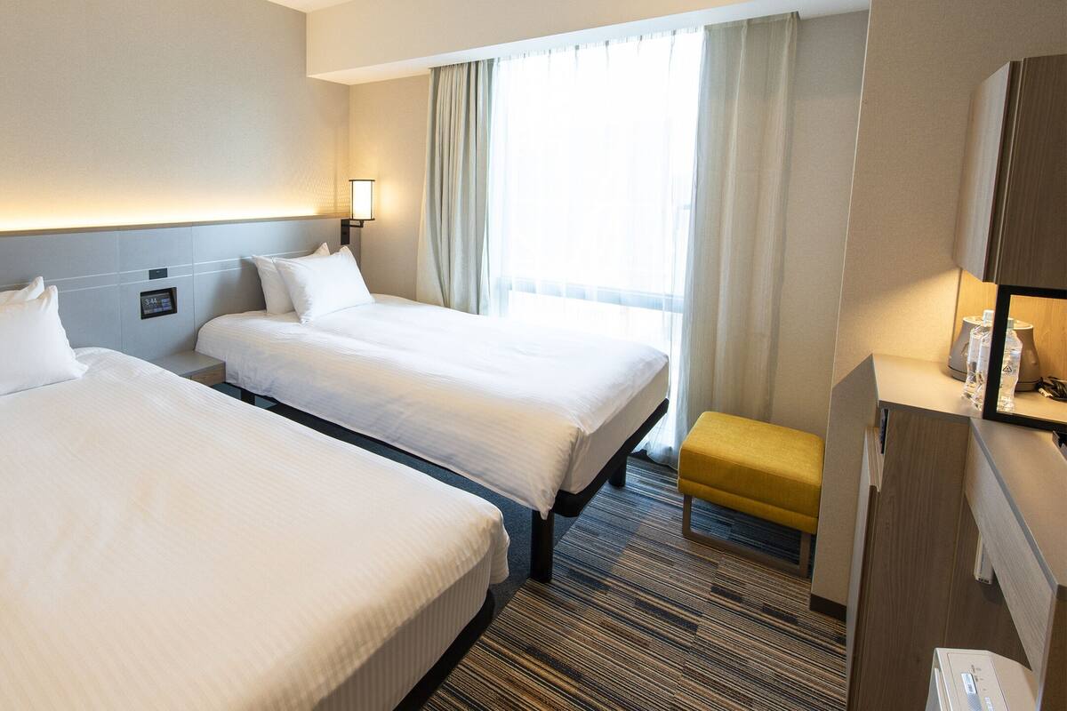 A hotel room with two beds, a desk, a chair, and a window with curtains.