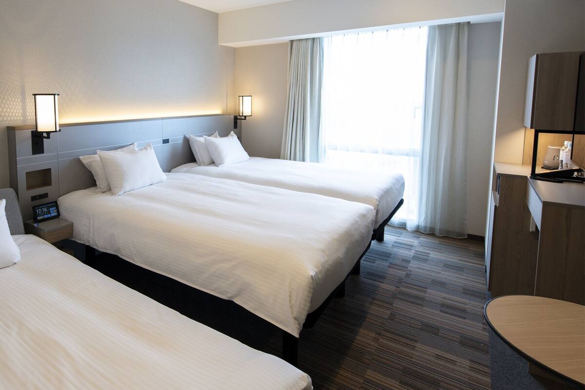 A hotel room with three beds, a bedside table with a clock, and a window with curtains.