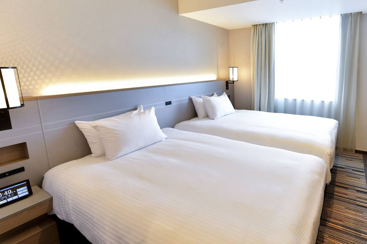 A hotel room with two beds, a TV, a bedside table with a clock, and a window with curtains.