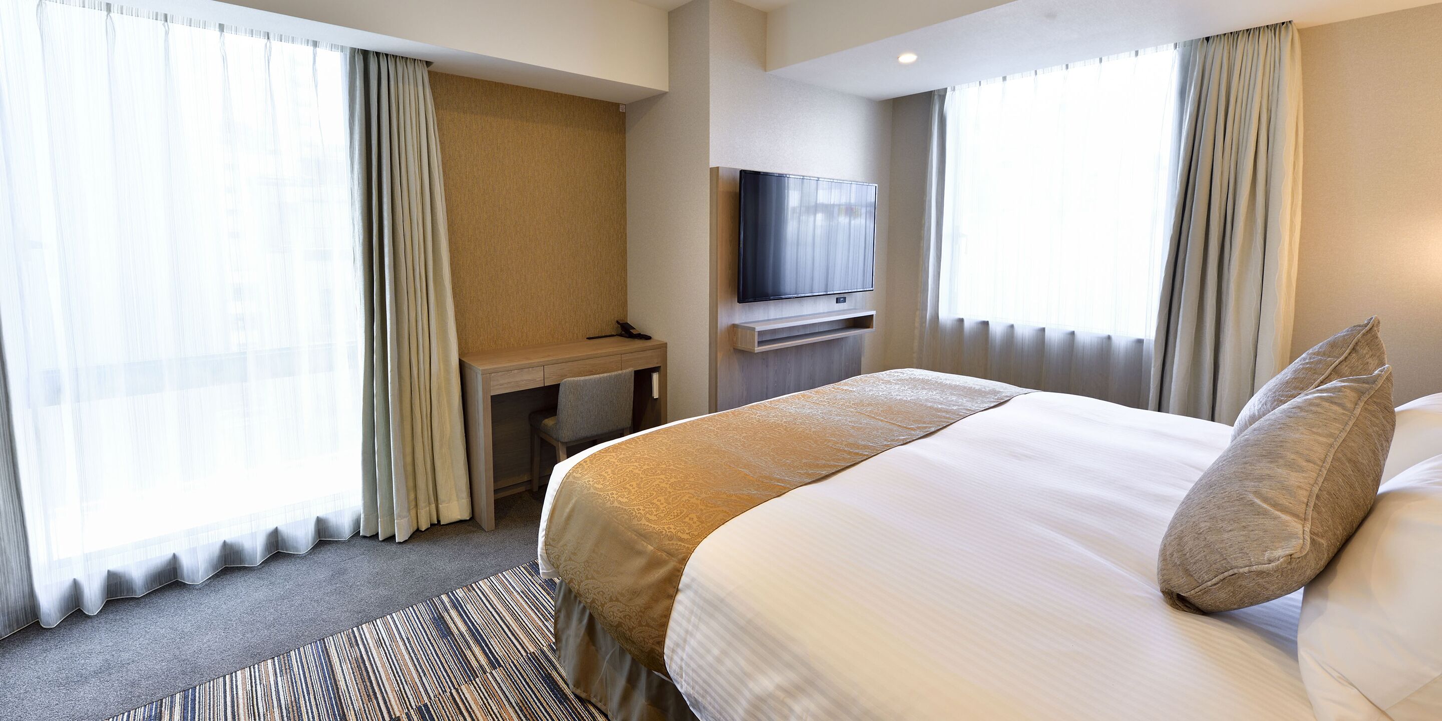 A hotel room with a bed, a desk, a television, and large windows with curtains.