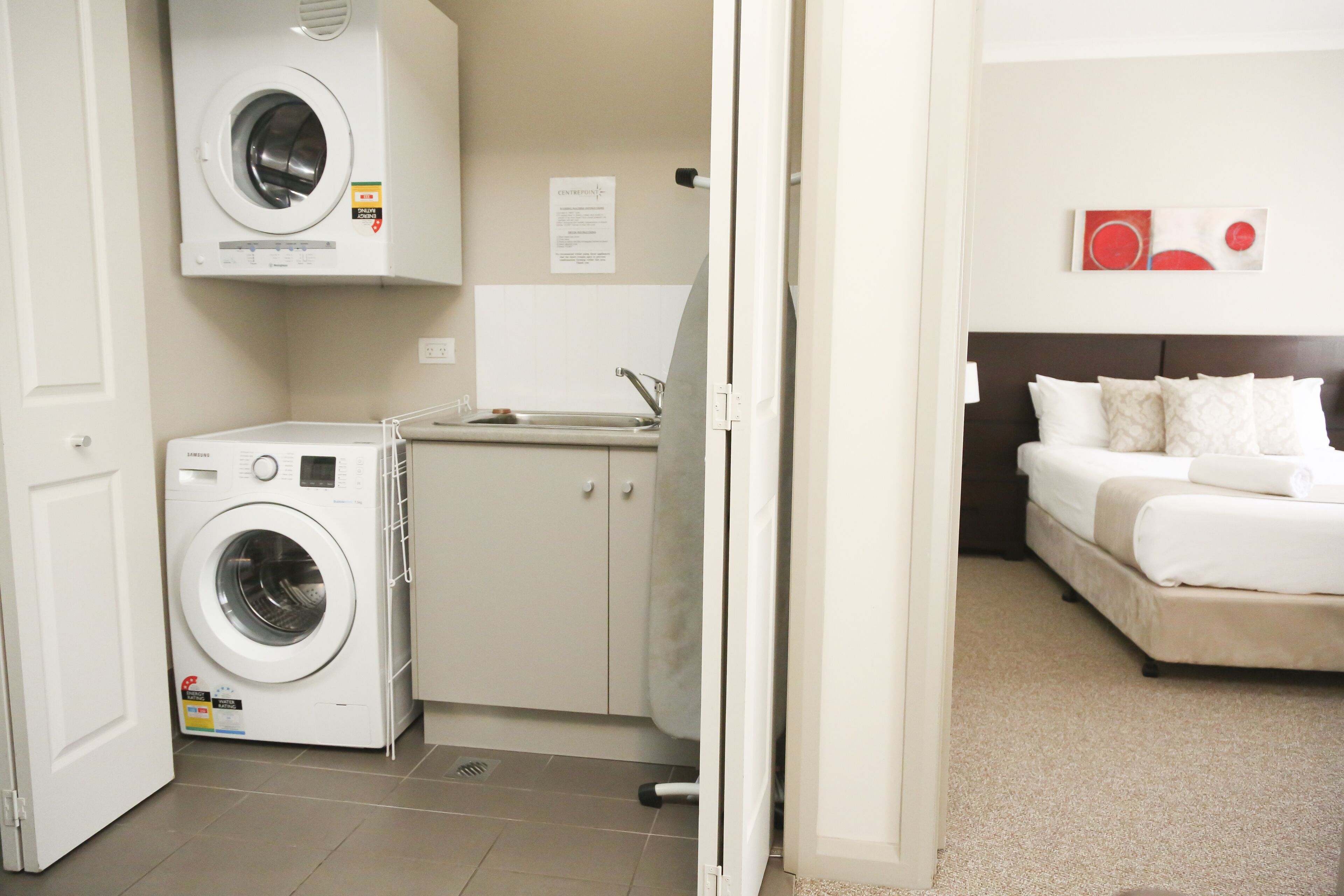 A compact hotel room with a washing machine, a small sink, and a bed with white bedding.